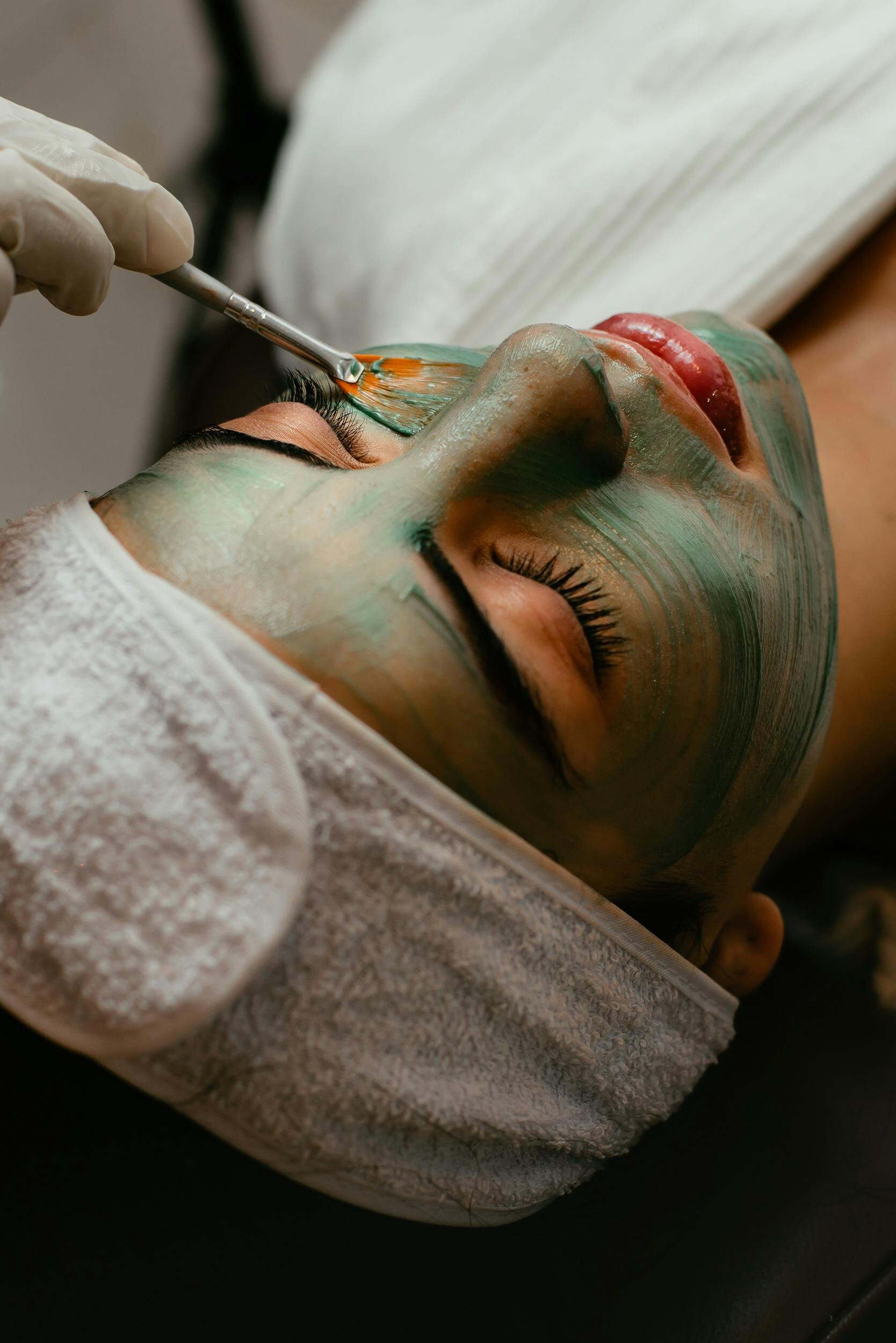 A woman relaxing while a green facial mask is applied with a brush