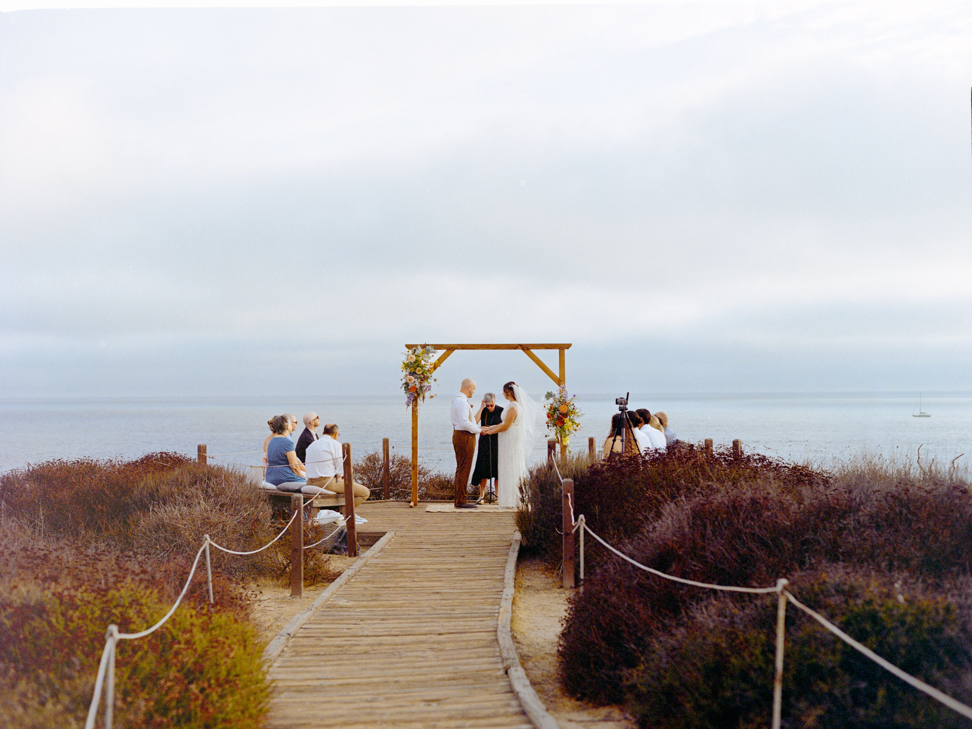 small elopement wedding ceremony on a cloudy overcast day at the beach overlooking the ocean. A bride and groom stand hand in hand under a wooden square arch with colorful flowers attached. Ten guests sit in a circle around the couple on a wooden boardwalk.
