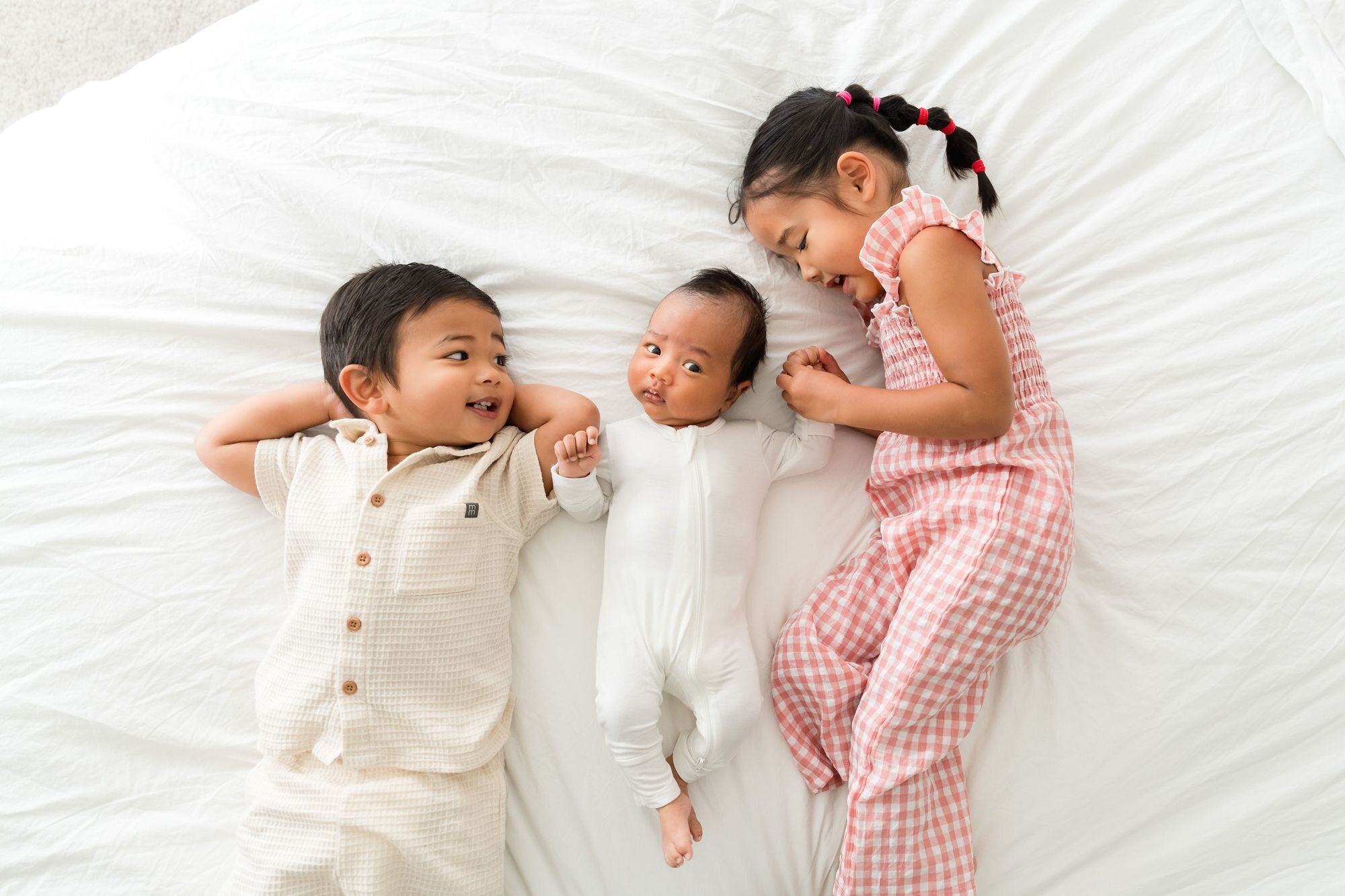 Big brother and big sister lying on the bed holding hands with their newborn sibling