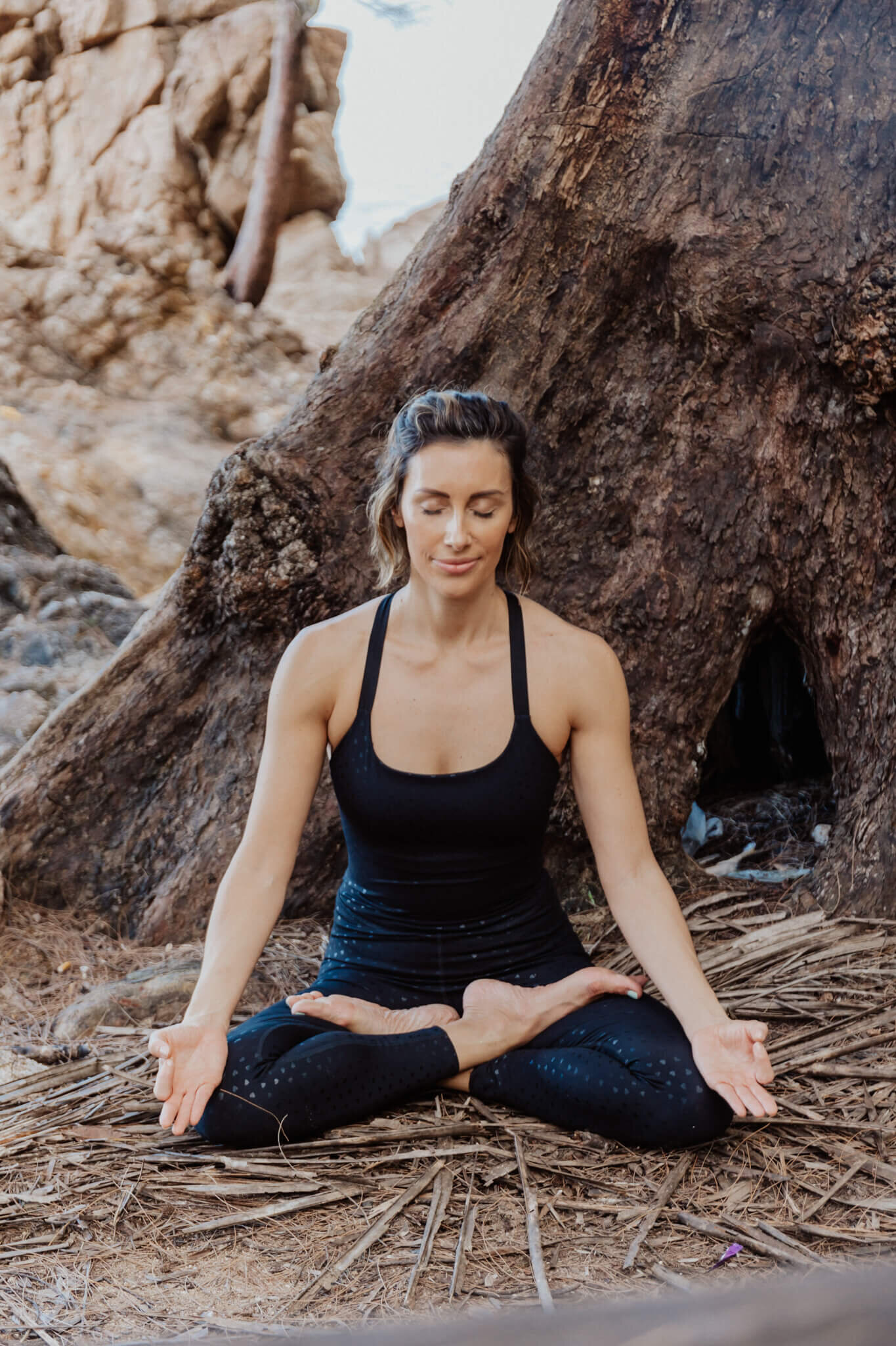 A woman practicing meditation in a seated yoga pose against a large tree trunk, representing the serene experience offered by our online yoga studio in Cape Town.