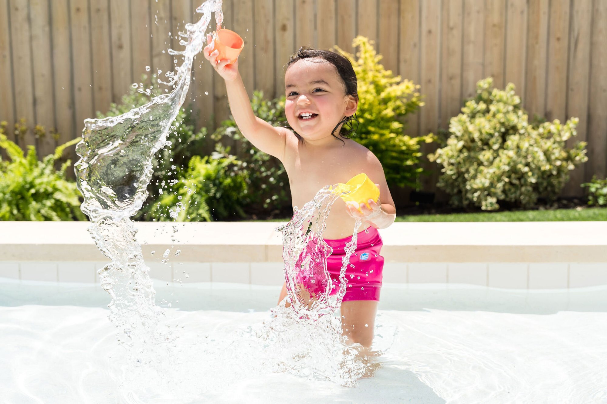 Toddler girl splashing in a backyard pool wearing Splash About swimwear, Austin