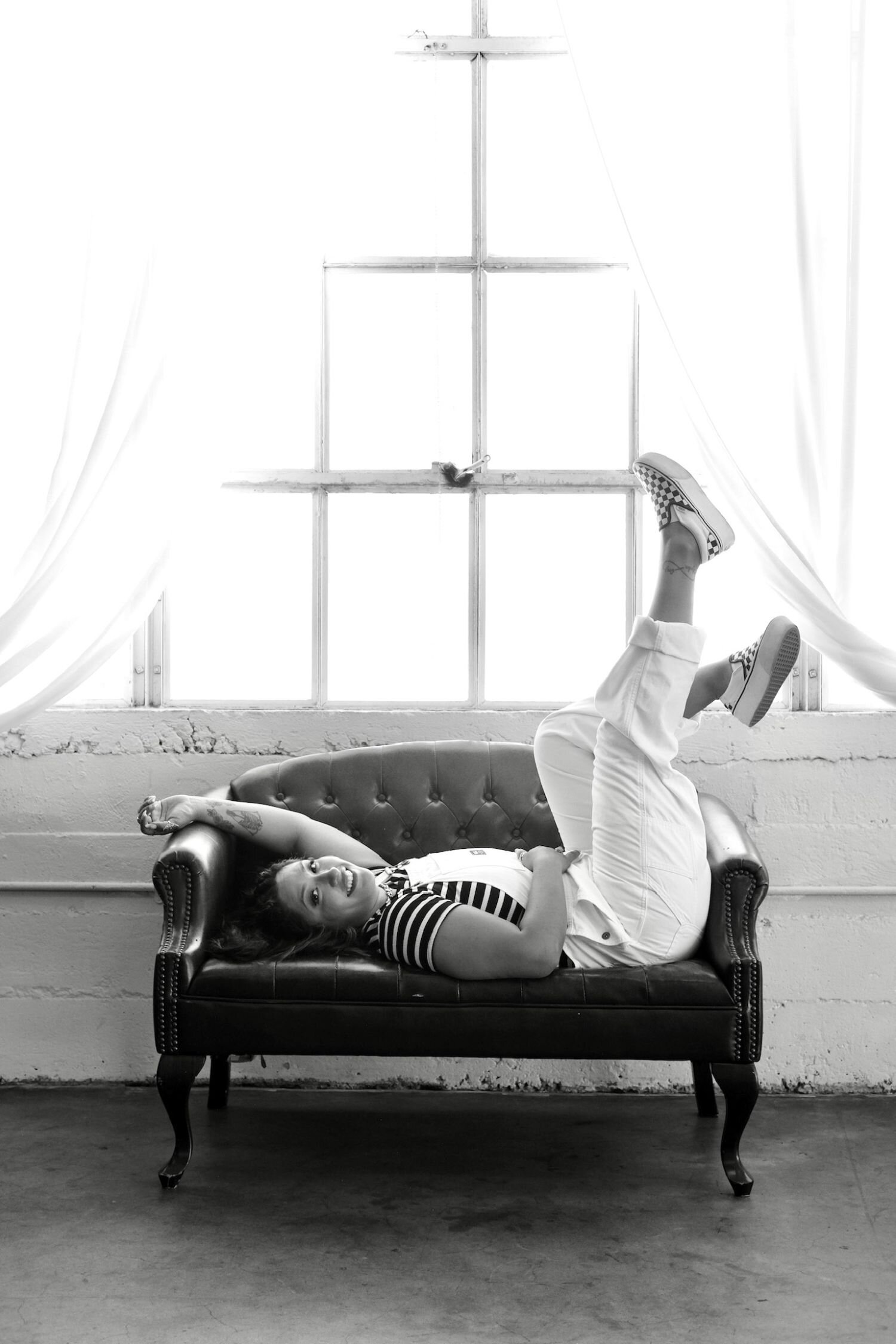 Joyce sitting on a brown leather chair, eyes closed and smiling with her tongue slightly out. She’s wearing a silver choker and hoop earrings, a sheer black top, and ripped jeans. Joyce is holding an open black notebook, her long almond-shaped nails painted white and stacked with silver rings. Behind her, a wooden wall features a large illuminated star-shaped marquee light.