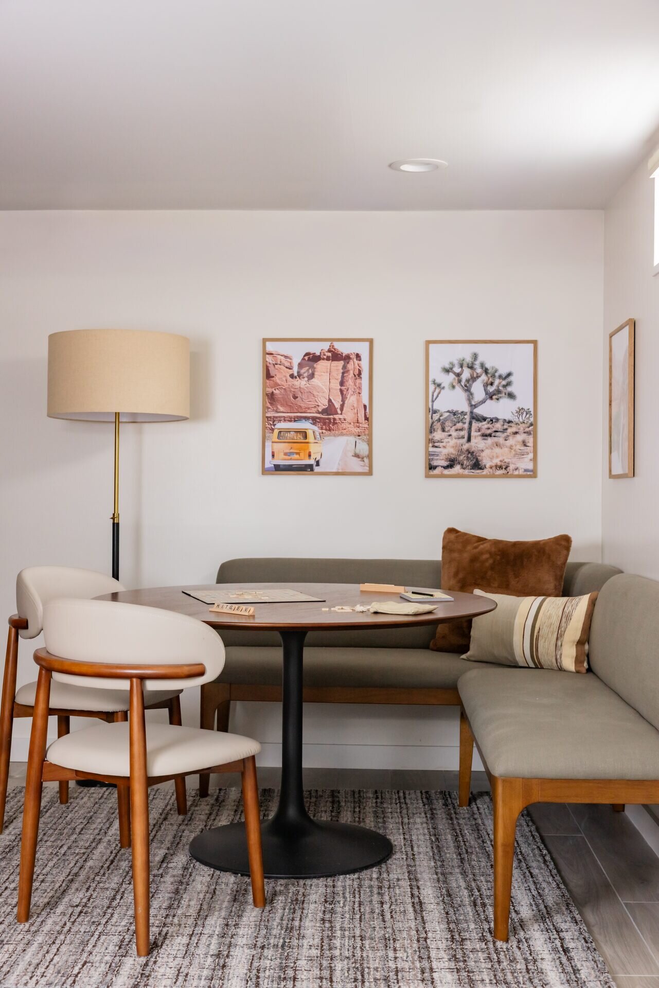 Basement dining nook with a round pedestal table, upholstered chairs, a corner bench with pillows, framed desert prints, and a floor lamp
