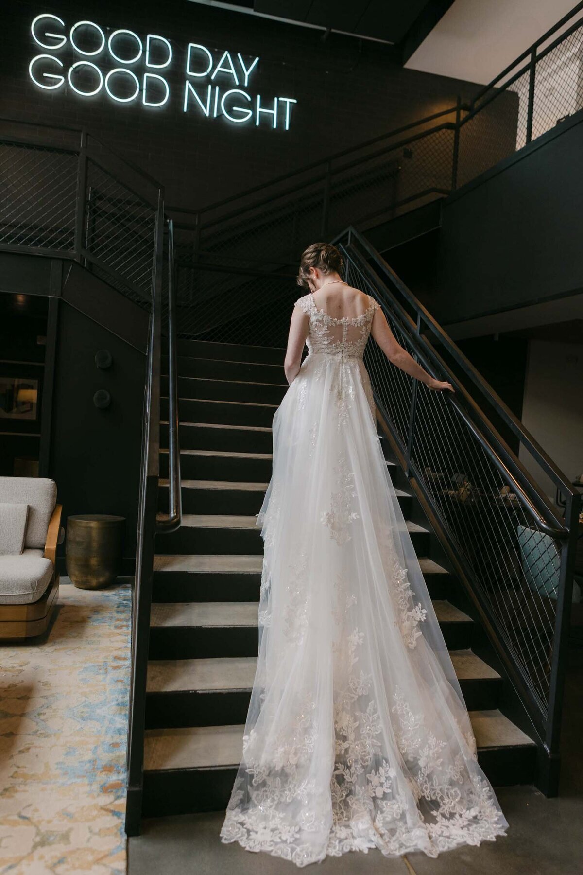 Bride with a lace train climbing a dark staircase beneath a Good Day Good Night neon sign