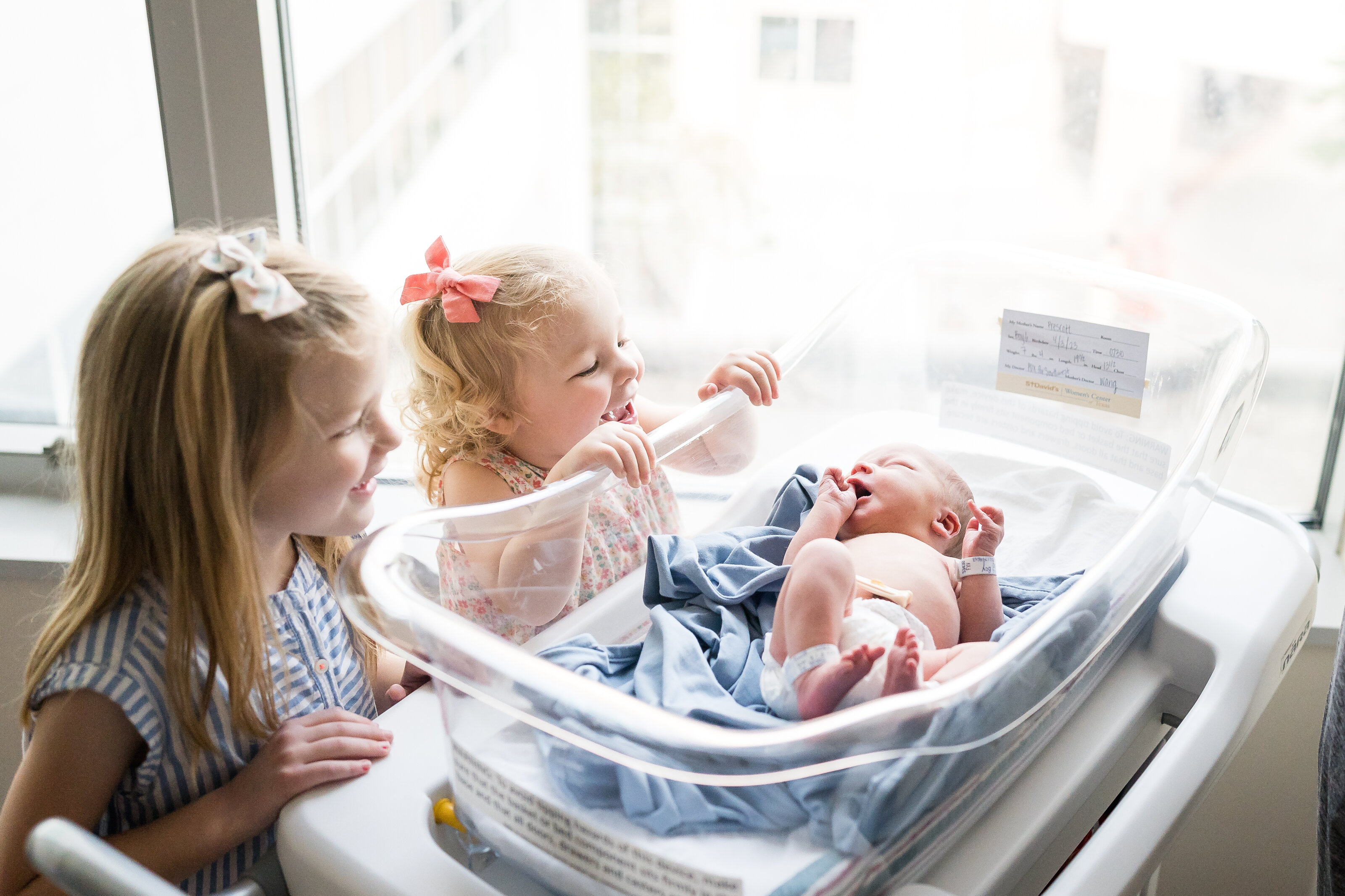 Two big sisters laughing as they meet their newborn baby brother in a hospital bassinet in Austin