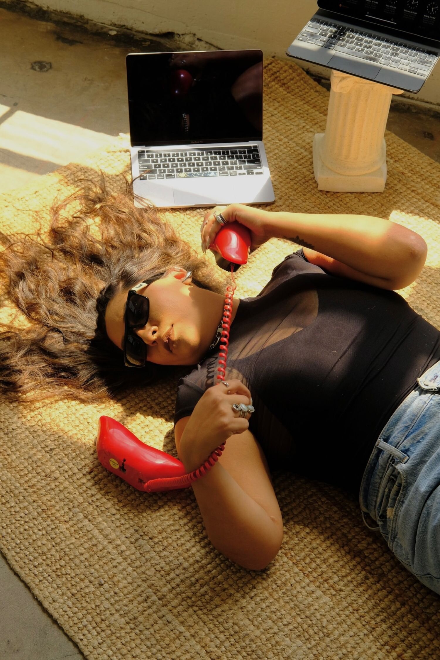 Joyce sitting on a brown leather chair, eyes closed and smiling with her tongue slightly out. She’s wearing a silver choker and hoop earrings, a sheer black top, and ripped jeans. Joyce is holding an open black notebook, her long almond-shaped nails painted white and stacked with silver rings. Behind her, a wooden wall features a large illuminated star-shaped marquee light.