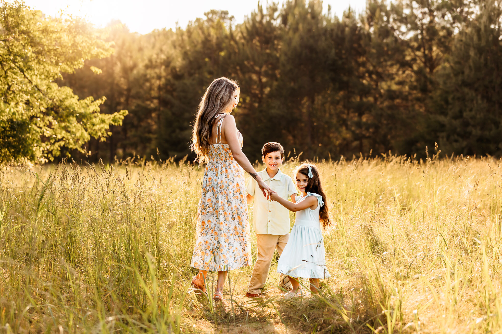 Mom in a floral maxi dress playing with her two kids, dressed in soft neutrals, in a sunny field