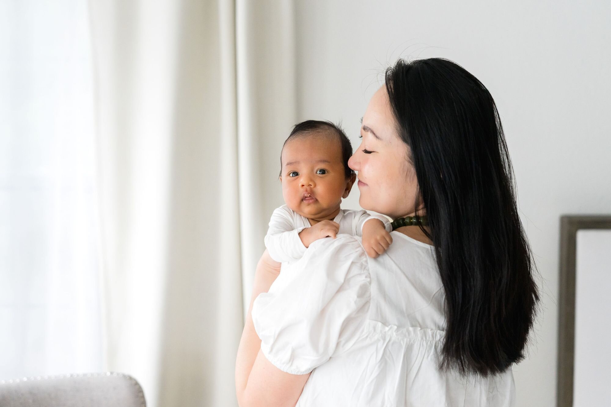 Mom holding her baby over her shoulder by a bright window at home
