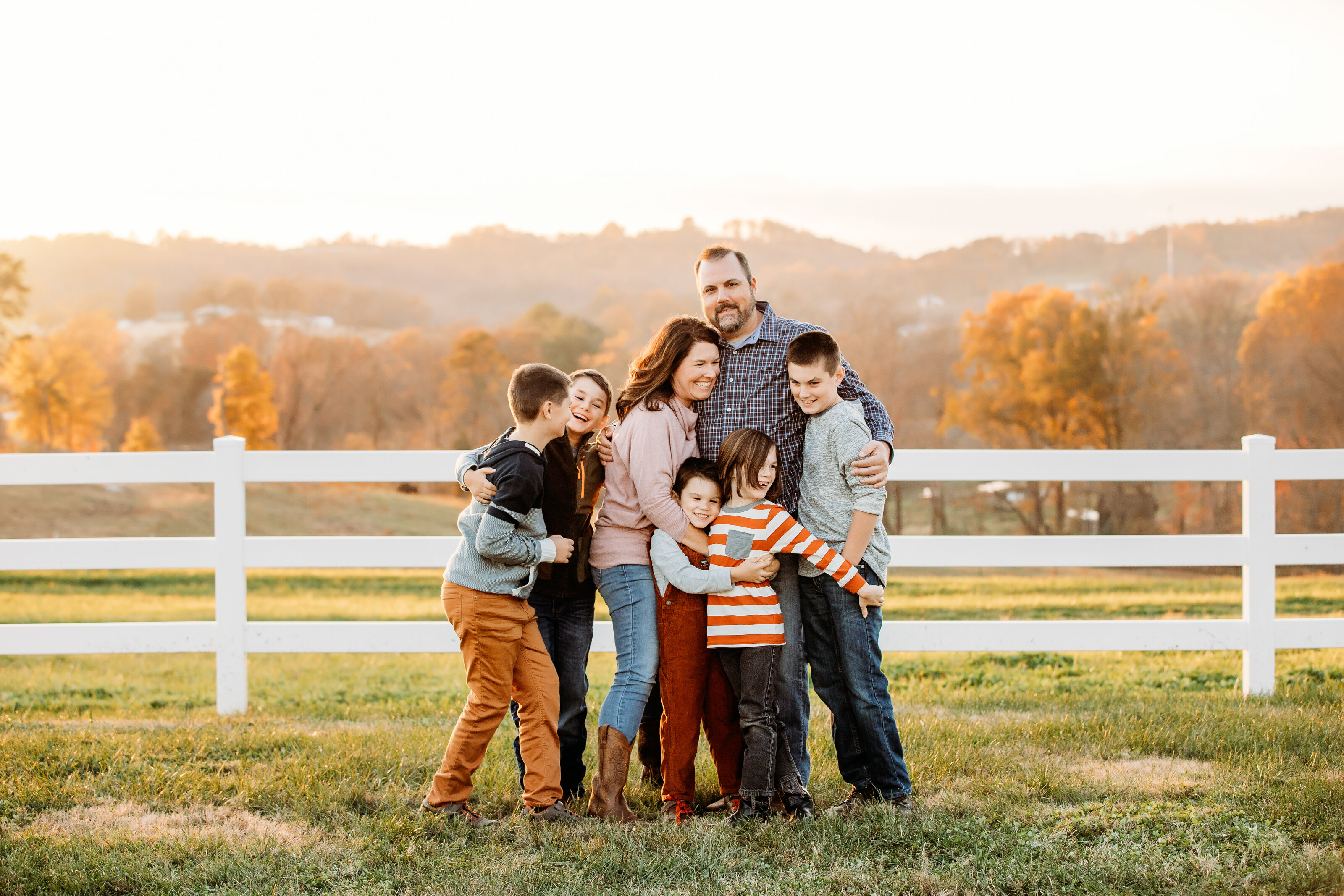 Family of six in coordinating fall tones hugging in front of a white fence