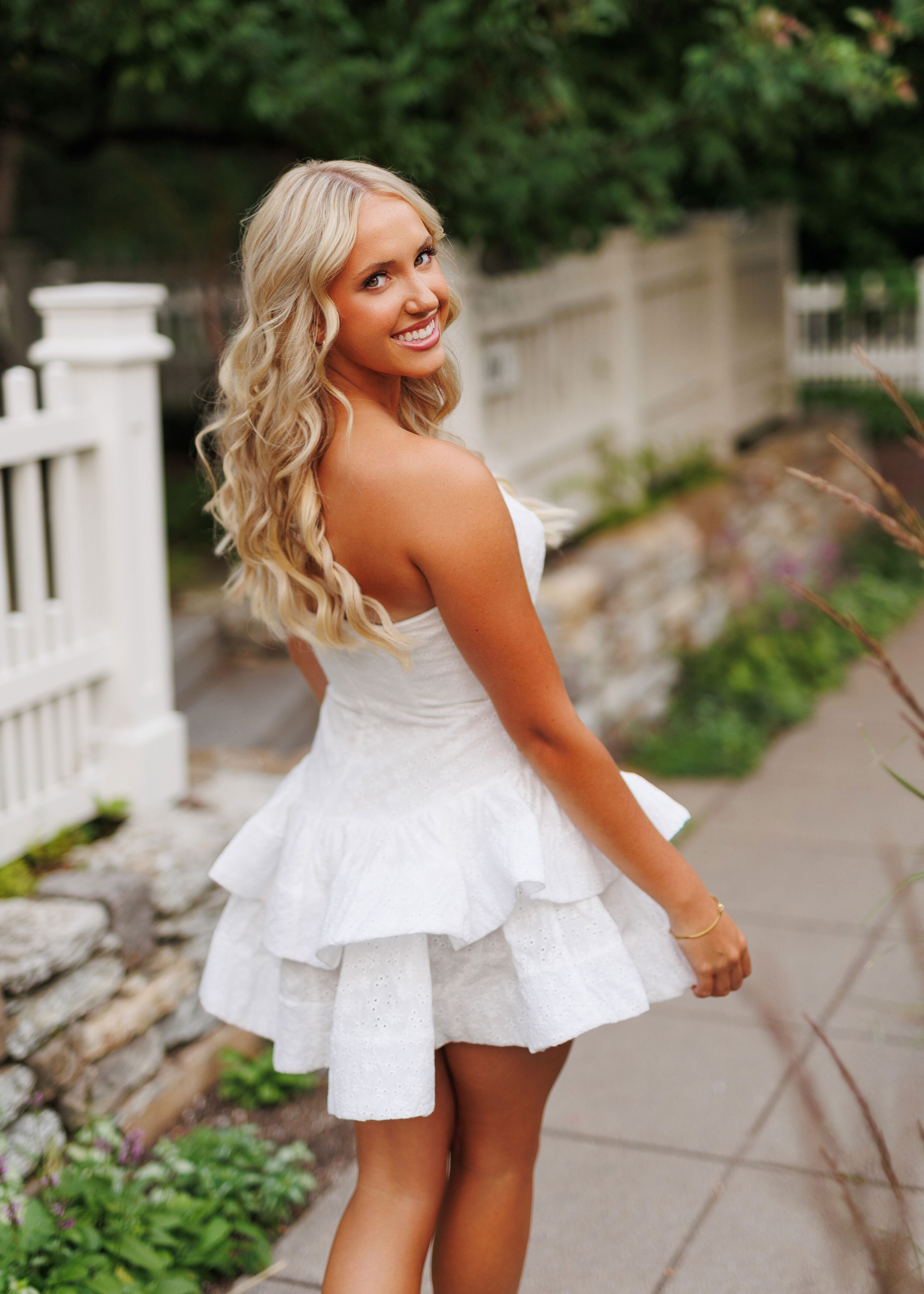 girl in white dress walking down flower lined minneapolis sidewalk during her summer senior photo session
