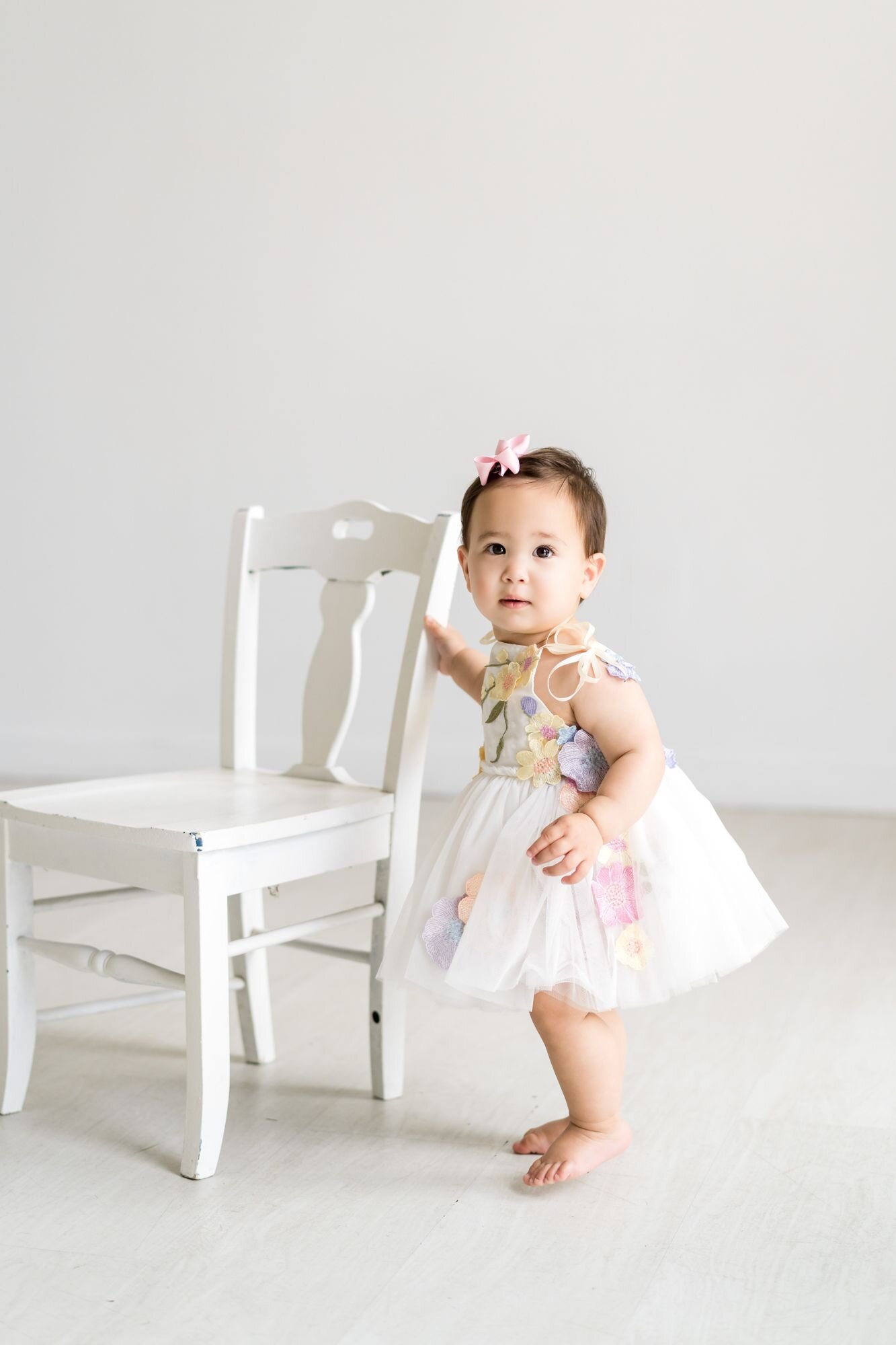 One-year-old girl in a floral tulle dress standing beside a white chair for her first birthday photos