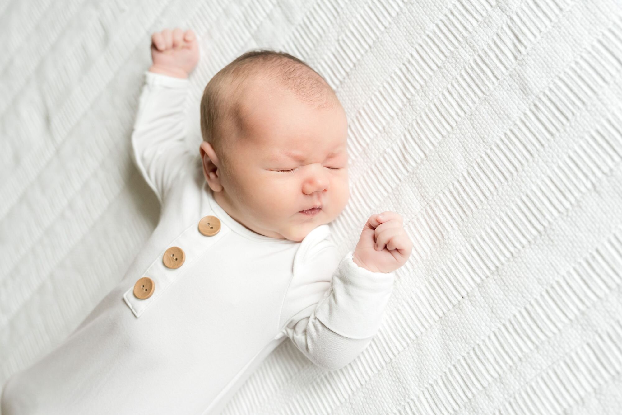 Newborn baby in a white button sleeper stretching on the bed during an in-home session in Austin