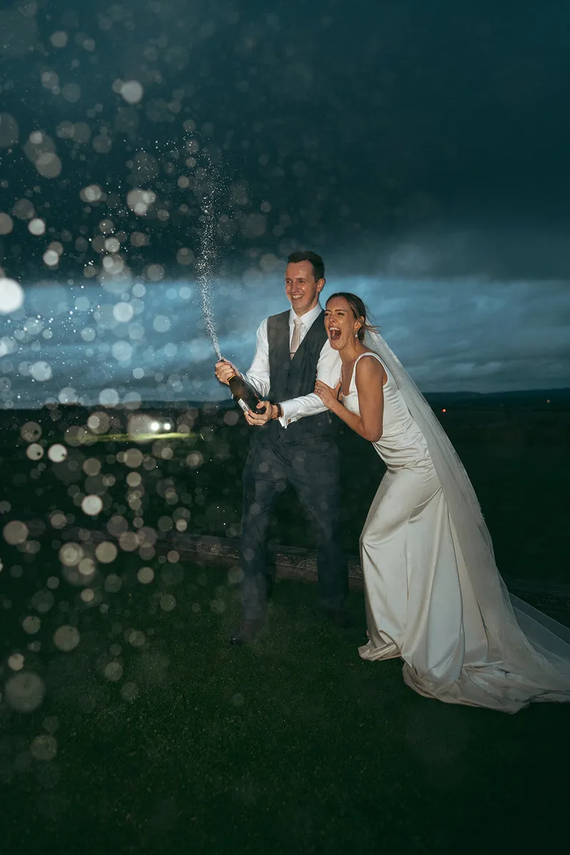 Groom spraying champagne over the bride under a stormy evening sky