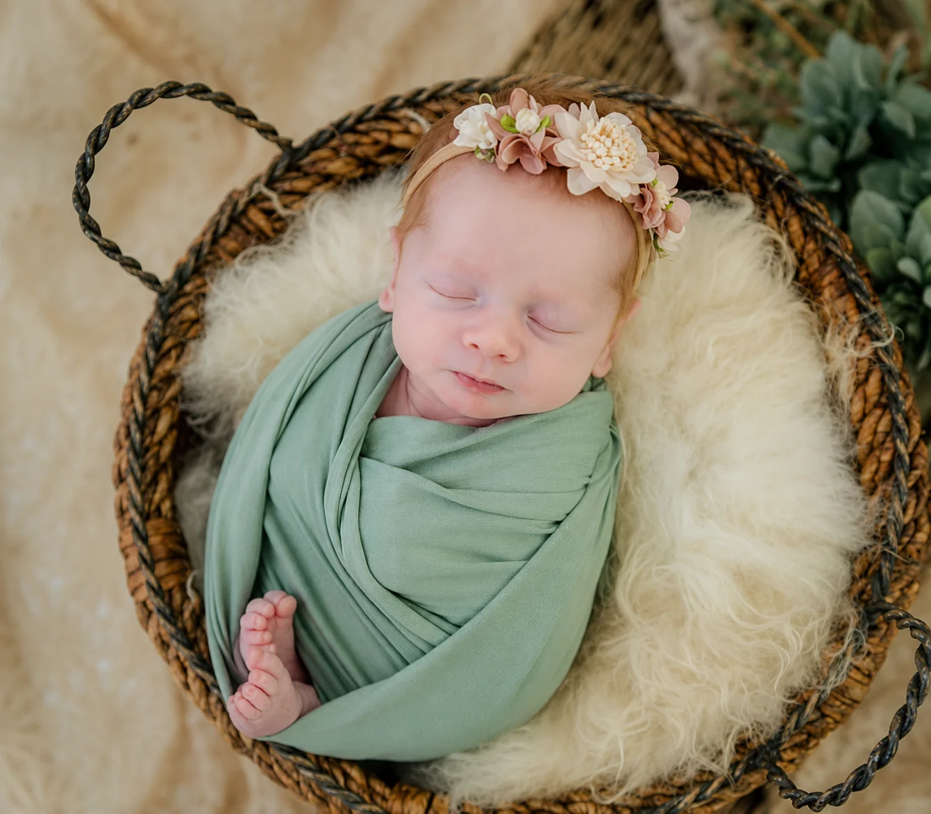 Newborn in a sage wrap and floral headband, resting in a woven basket